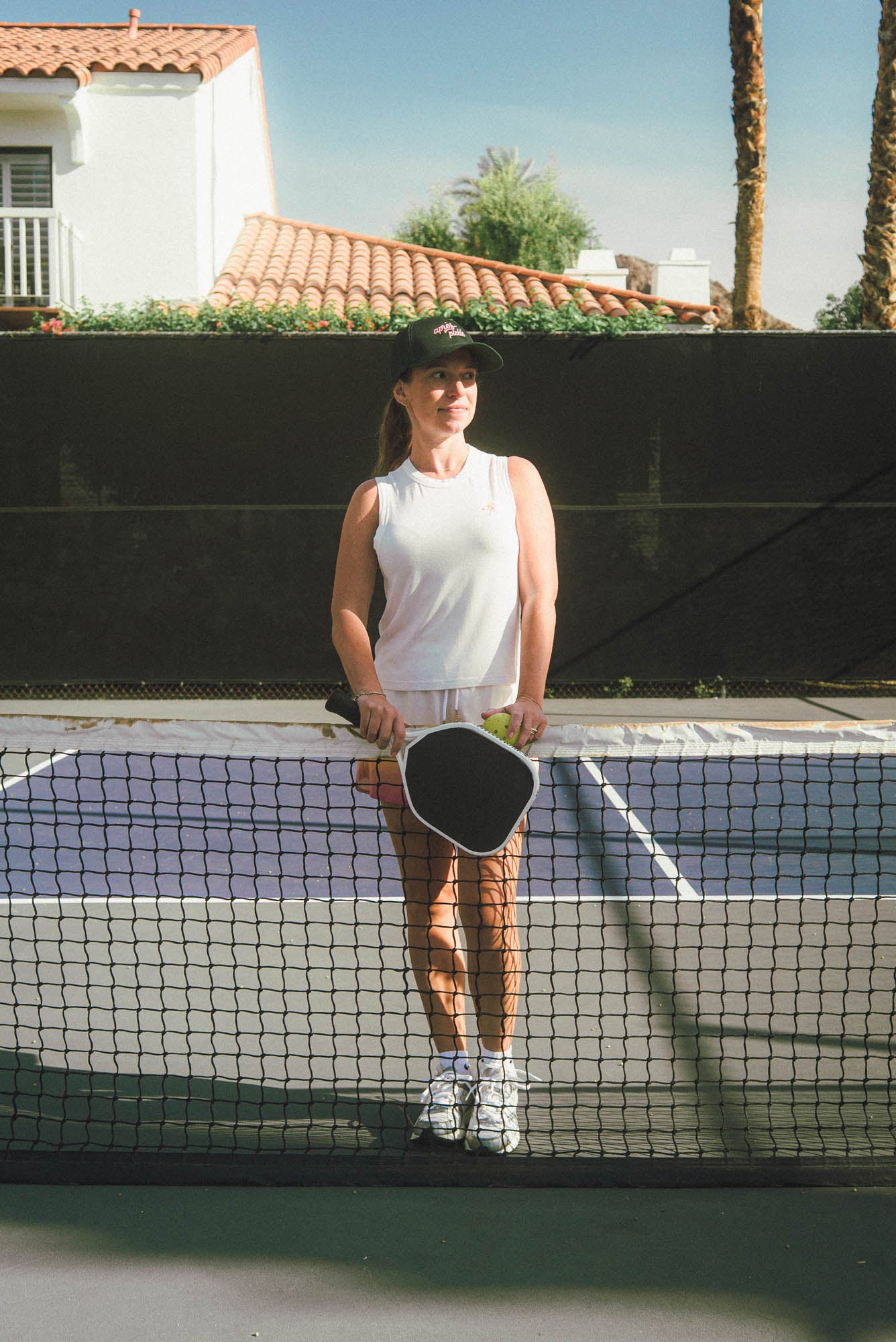 Woman on a tennis court holding a paddle and ball, with a residential area in the background.